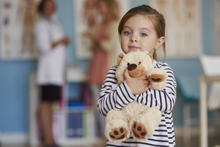 A little girl facing the camera holding a stuffed teddy while her mom watches in the background.